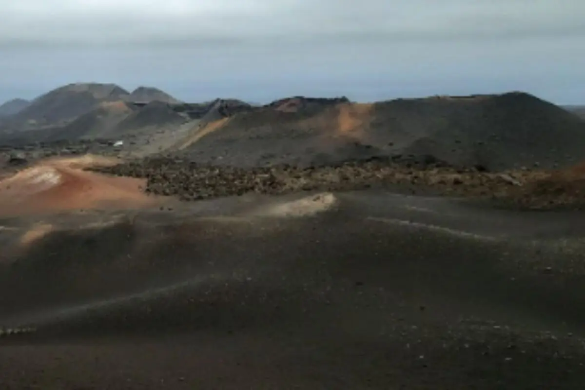 Paisatge volcànic de Lanzarote amb turons foscos de lava solidificada i tons vermellosos sota un cel ennuvolat.