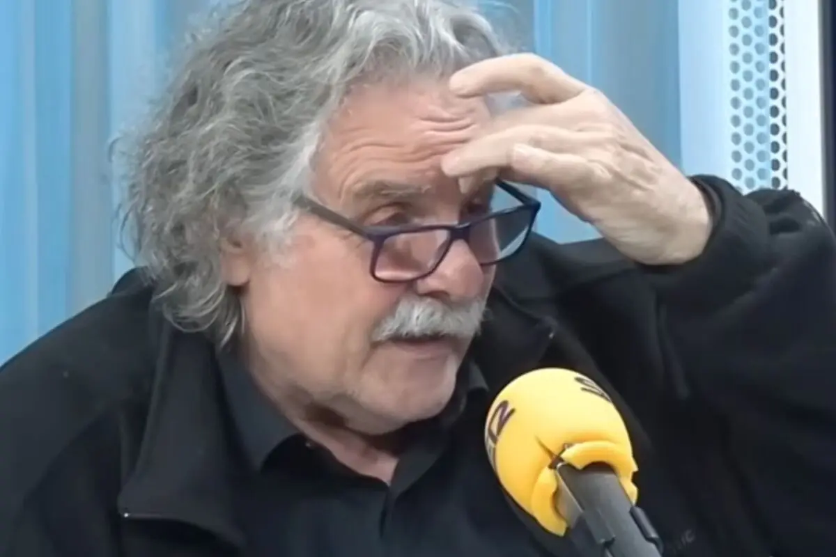 Hombre mayor con gafas y bigote canoso se toca la frente mientras habla frente a un micrófono amarillo en un estudio de radio