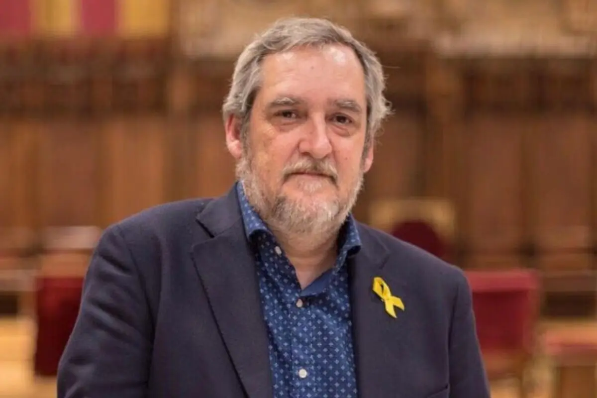 Home de cabells canosos i barba amb jaqueta blava i camisa blava amb petits punts blancs porta un llaç groc a la solapa i es troba en un interior elegant amb el fons desenfocat