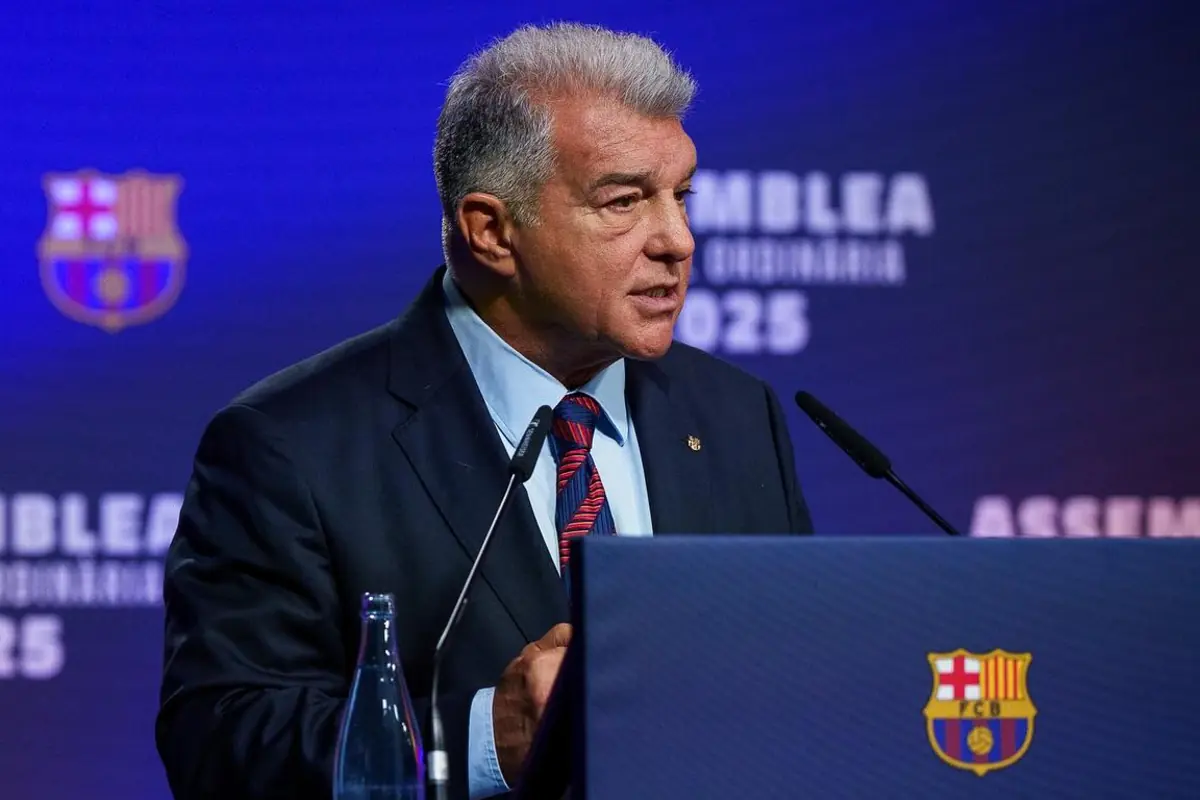 Gray-haired man in a suit and tie speaking at a podium with the FC Barcelona crest and a blue background with blurred text