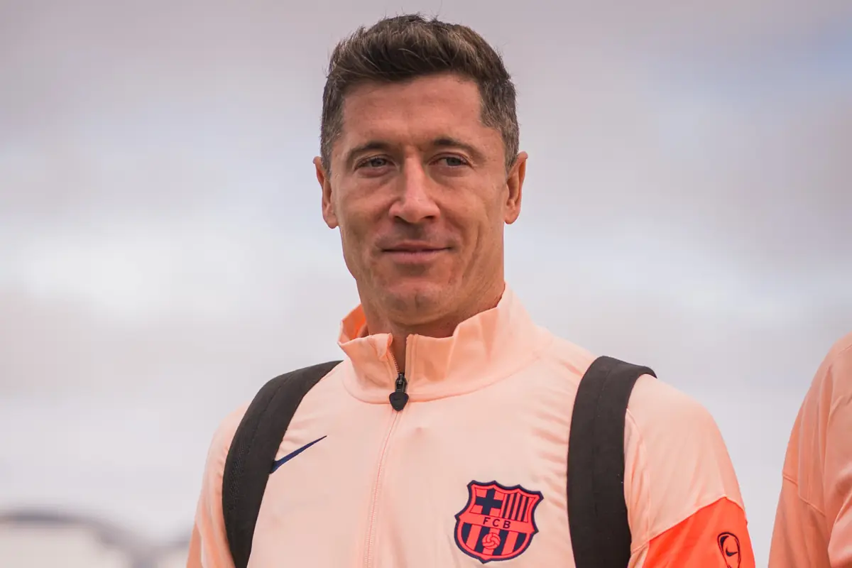 Home amb jaqueta esportiva de color préssec i escut del FC Barcelona, porta motxilla i és a l’aire lliure