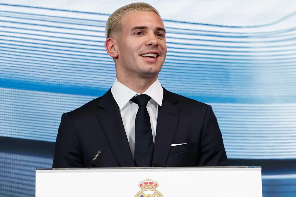 Young man with short blond hair dressed in a dark suit and black tie speaking at a podium with the Real Madrid crest on the front and a light blue background.