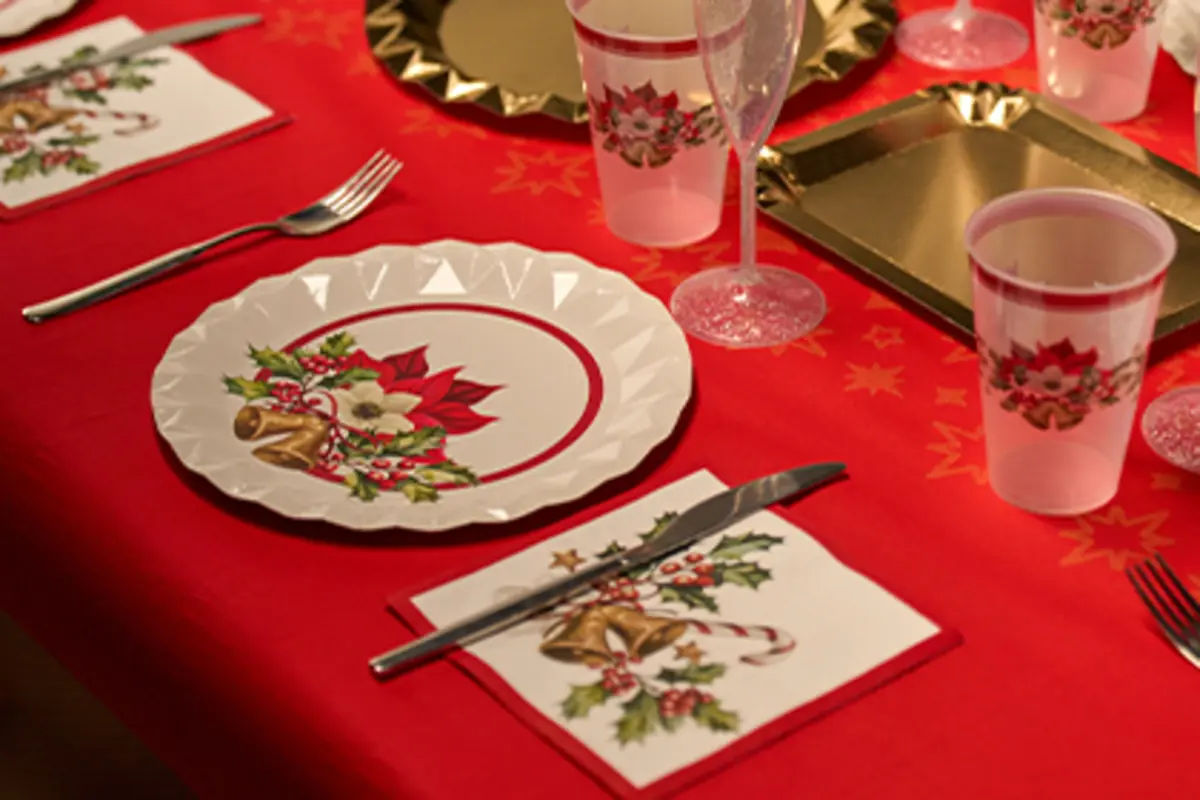 Mesa decorada con vajilla desechable de temática navideña sobre mantel rojo, con platos, vasos, copas y servilletas adornadas con flores de pascua y campanas Mesa decorada con vajilla desechable de temática navideña sobre mantel rojo, con platos, vasos, copas y servilletas adornadas con flores de pascua y campanas