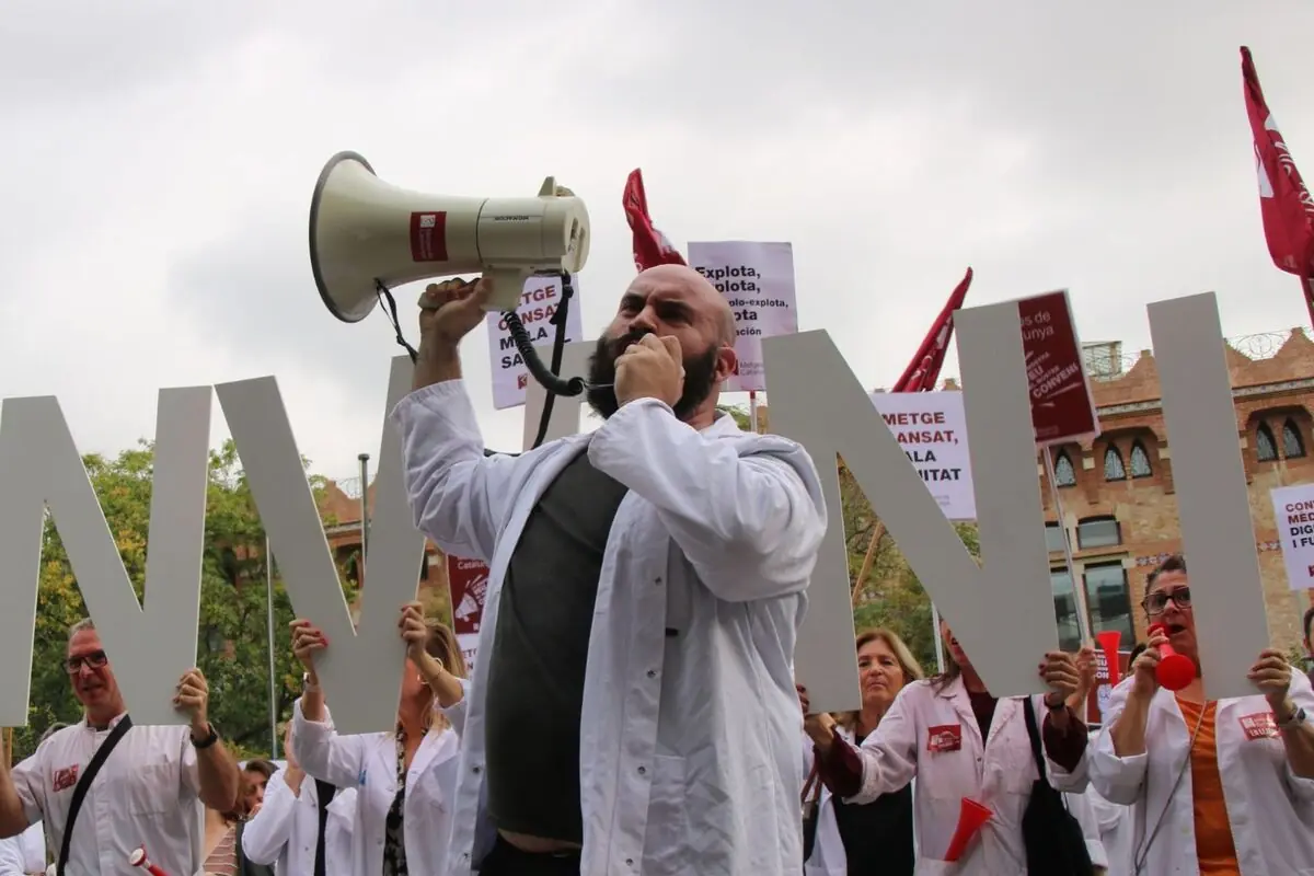 Un grup de persones amb bates blanques participa en una manifestació, una persona al davant sosté un megàfon i crida mentre altres aixequen lletres grans i pancartes.