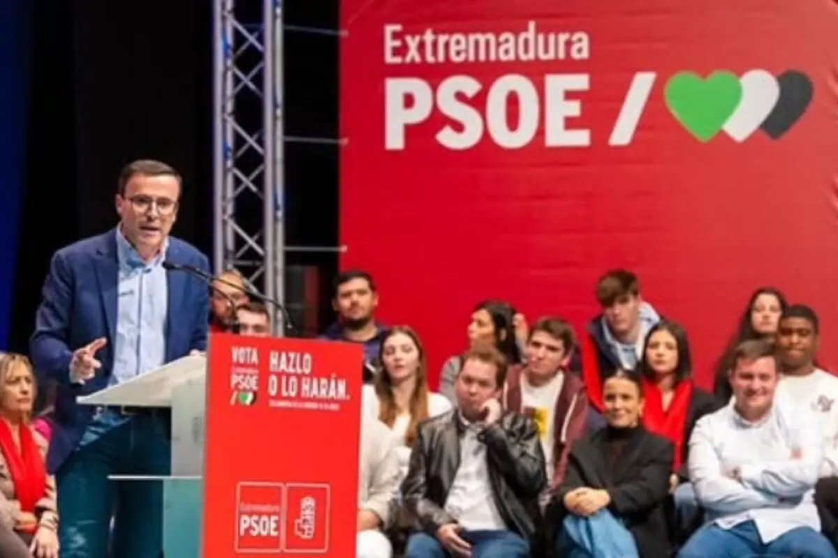 Miguel Ángel Gallardo Miranda fent un discurs en un míting del PSOE a Extremadura amb un gran cartell vermell de fons i diverses persones assegudes escoltant a l’escenari.