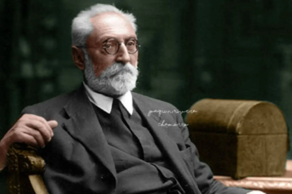 Retrat en color de Miguel de Unamuno amb barba i bigoti blancs assegut amb un vestit fosc i ulleres rodones davant d’un fons verd fosc.