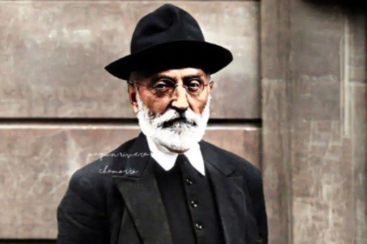 Miguel de Unamuno amb barba blanca, ulleres i barret negre posant amb un vestit fosc davant d’una paret de pedra.