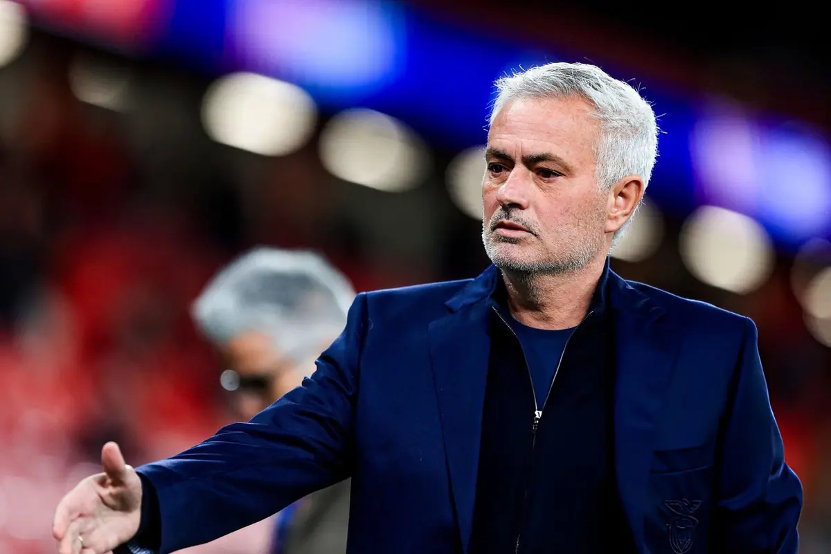 Home de cabells canosos amb barba curta, vestit amb una jaqueta blau fosc en un estadi il·luminat, estenent la mà