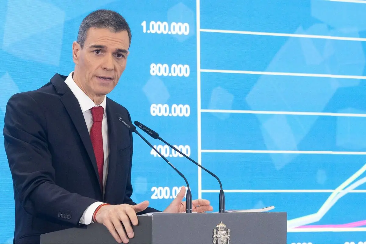 Hombre con traje oscuro y corbata roja hablando en un atril con micrófonos frente a una pantalla azul con un gráfico de líneas y cifras grandes en el fondo