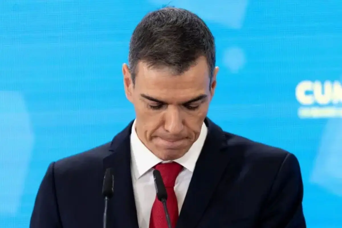 Hombre con traje oscuro y corbata roja mirando hacia abajo frente a un micrófono sobre fondo azul