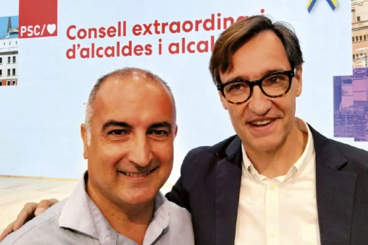 Dos hombres sonríen juntos frente a un cartel de un evento del PSC