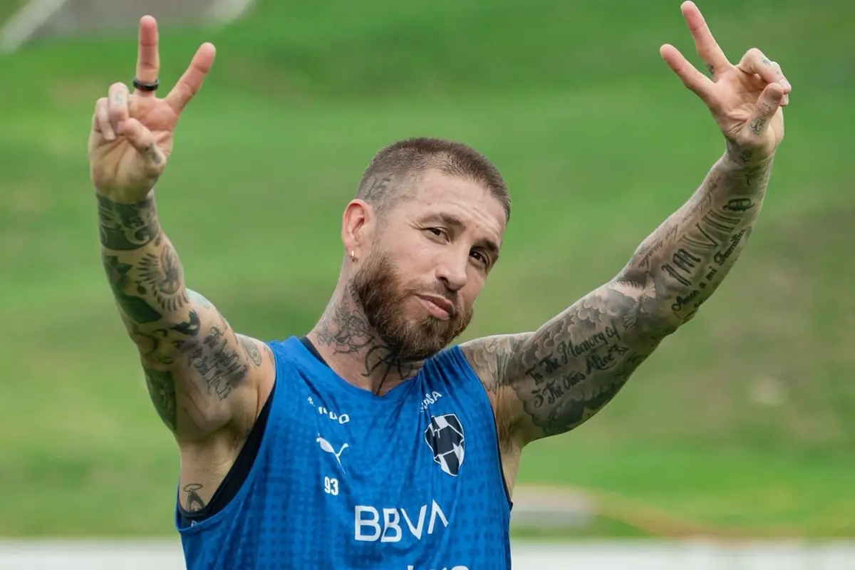 Bearded man with tattoos on his arms wearing a blue workout jersey raising both hands making the peace sign with his fingers on a grass field