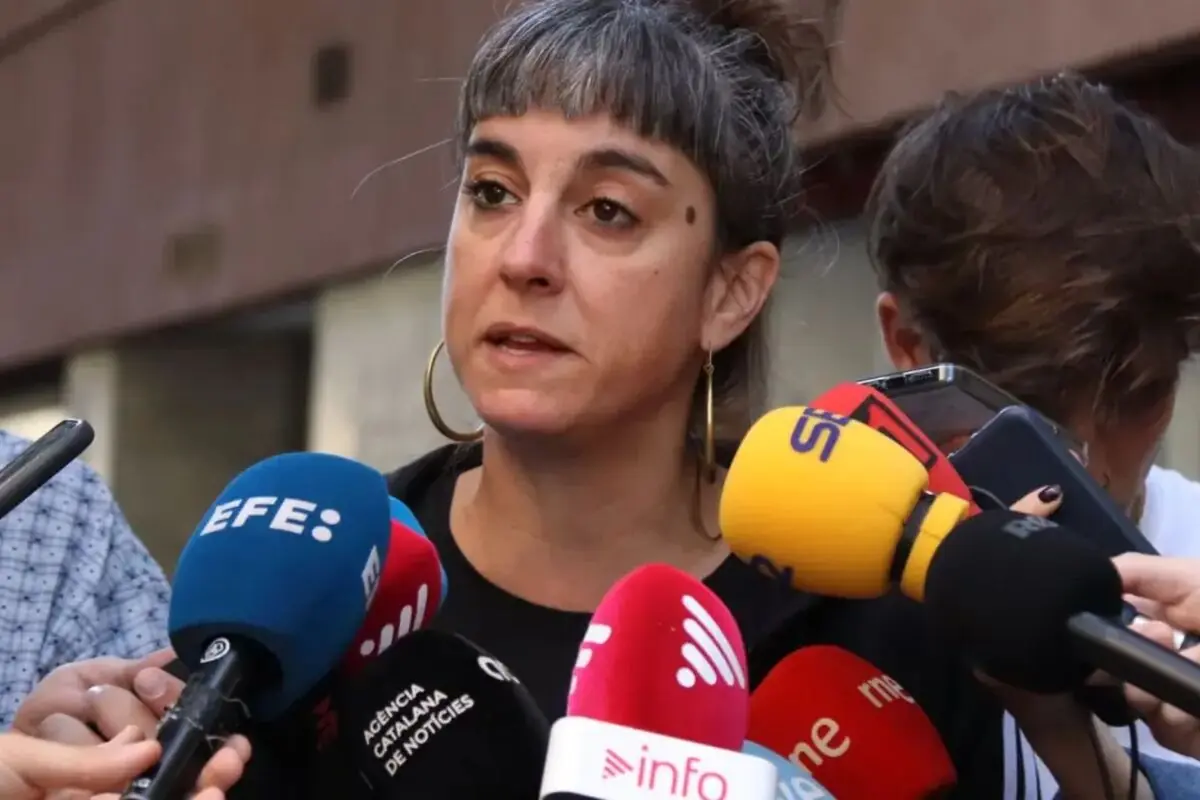 Mujer con moño y pendientes de aro hablando ante varios micrófonos de diferentes medios de comunicación en una rueda de prensa al aire libre