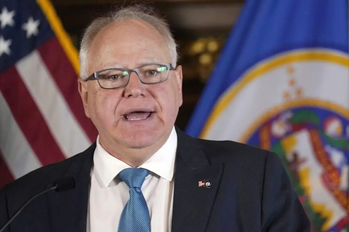 Hombre mayor con gafas y traje hablando frente a micrófono con banderas de Estados Unidos y de un estado estadounidense de fondo