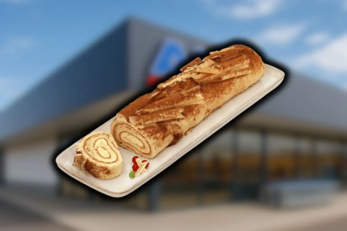 Brazo de gitano de Aldi relleno de crema presentado en una bandeja blanca con una rodaja cortada delante de un fondo desenfocado de supermercado.