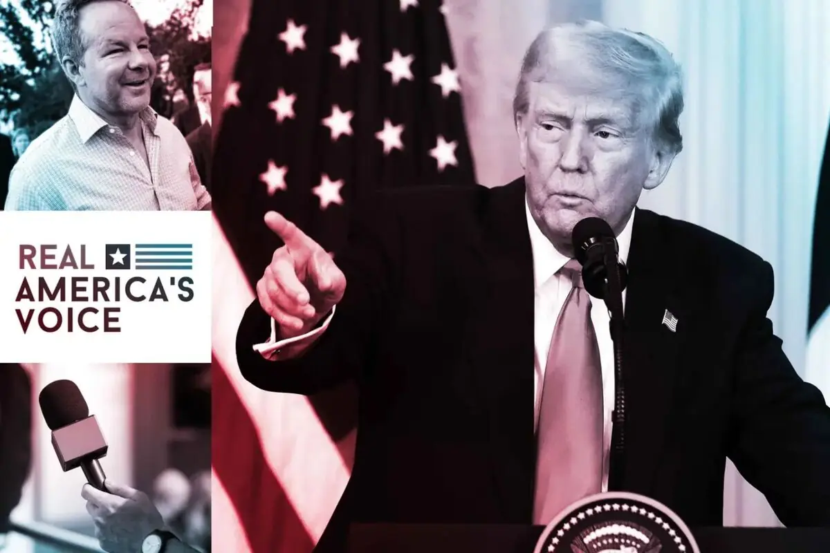 Un hombre de traje y corbata habla en un podio con micrófono mientras señala con el dedo, al fondo hay una bandera de Estados Unidos y en la esquina izquierda aparece el logo de Real America's Voice junto a un hombre sonriente y un micrófono de reportero.