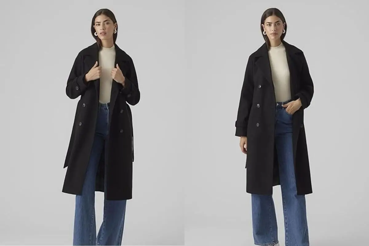 Mujer de pie con abrigo negro largo de doble botonadura suéter beige y vaqueros anchos azules sobre fondo gris claro