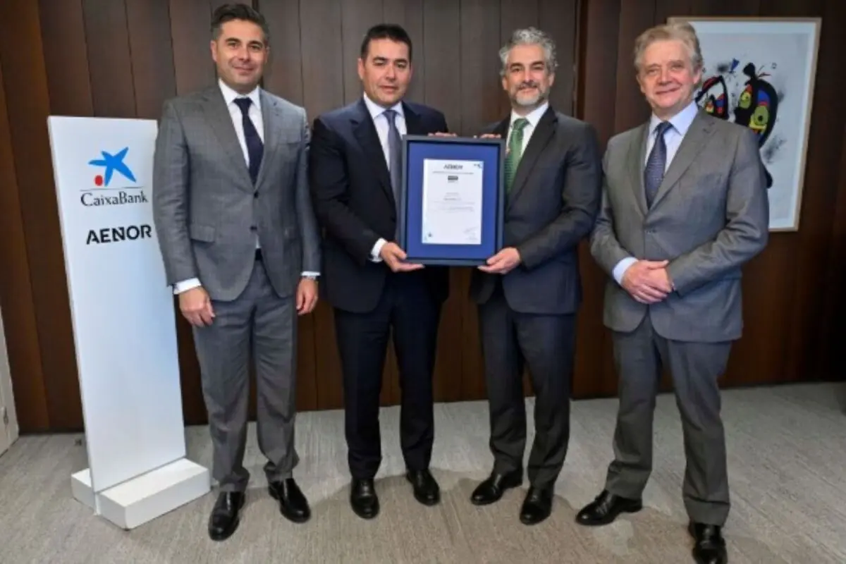 Quatre homes amb vestit posen en una oficina mentre dos d’ells sostenen un diploma emmarcat al costat d’un panell amb els logotips de CaixaBank i AENOR