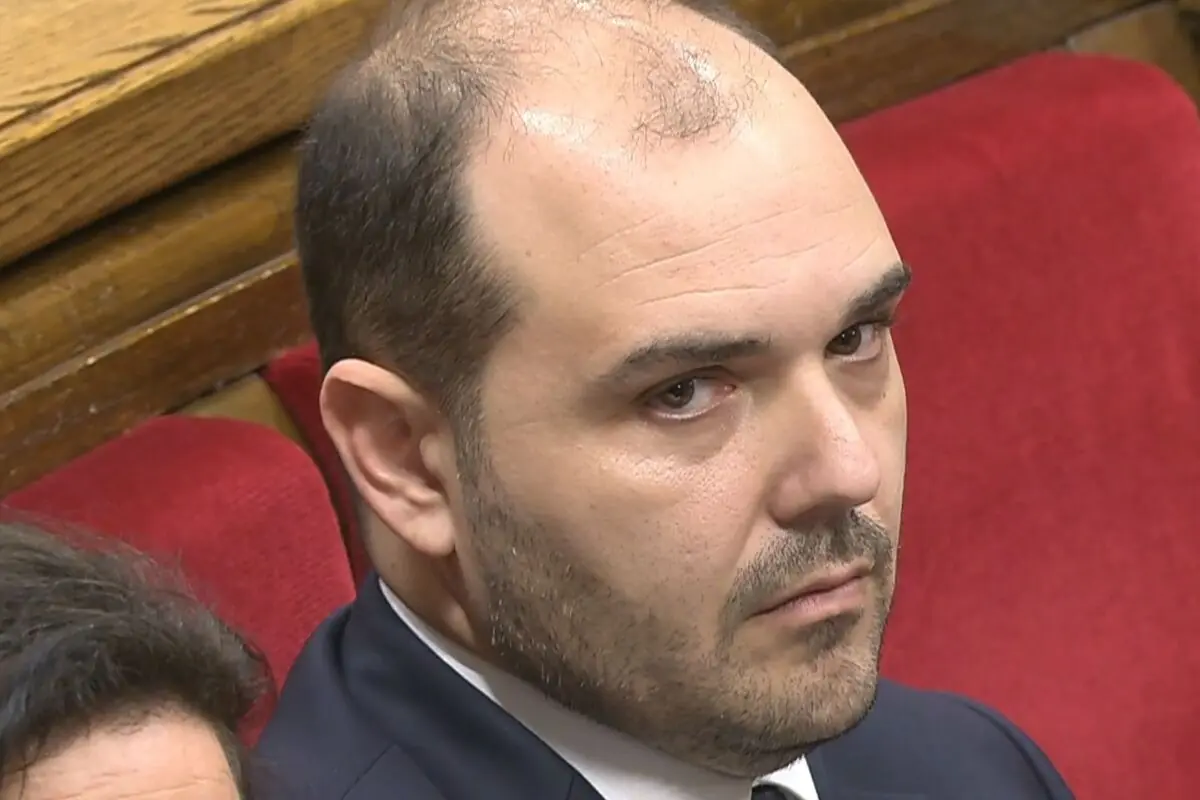 Home de mitjana edat amb vestit fosc assegut en un seient vermell mirant seriosament cap a la càmera