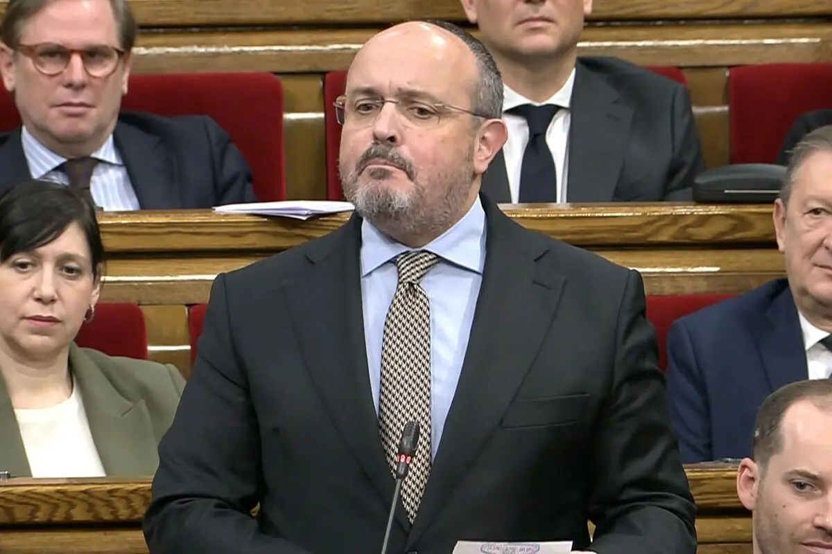 Hombre calvo con barba y traje oscuro hablando frente a un micrófono en un parlamento lleno de personas sentadas en escaños de madera con asientos rojos