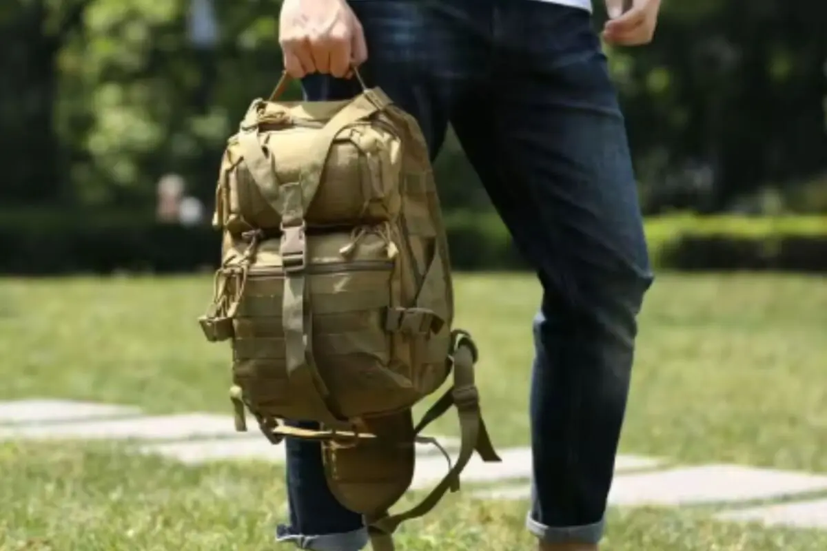 Persona de pie en un parque sosteniendo con la mano una mochila táctica color caqui Persona de pie en un parque sosteniendo con la mano una mochila táctica color caqui