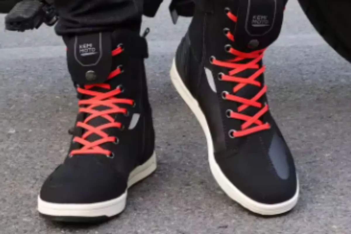 Botas negras de moto tipo zapatilla con cordones rojos y suela blanca sobre pavimento gris