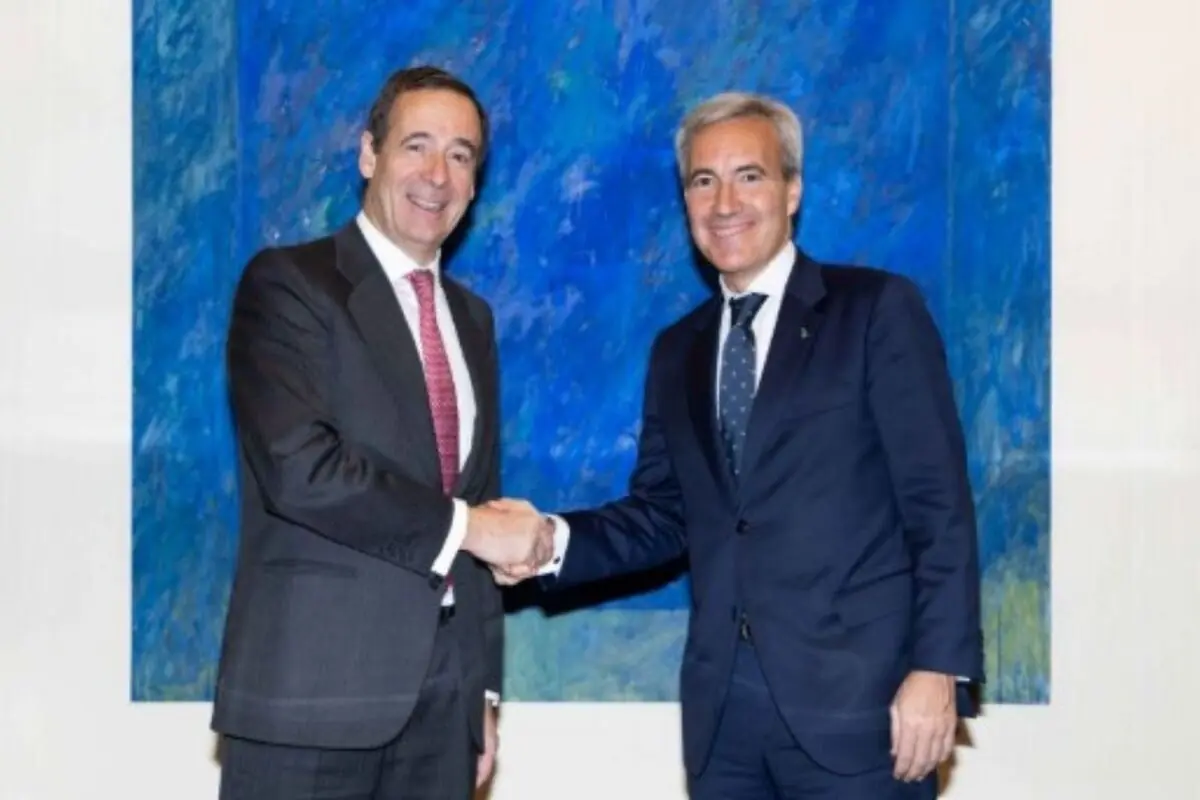 Dos homes de negocis amb vestit es donen la mà somrient davant d’un gran quadre blau