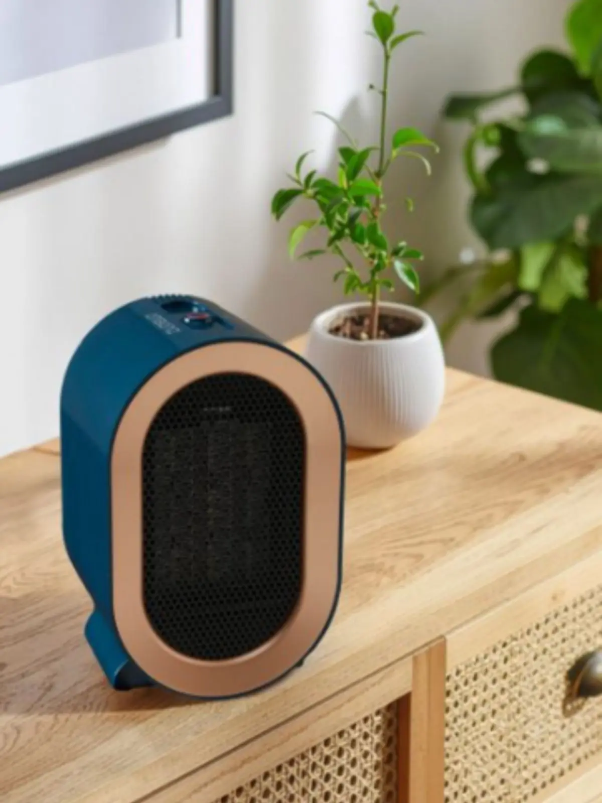 Calefactor eléctrico compacto de color azul y dorado de Aldi sobre una cómoda de madera junto a una maceta con planta verde. Calefactor eléctrico compacto de color azul y dorado de Aldi sobre una cómoda de madera junto a una maceta con planta verde.