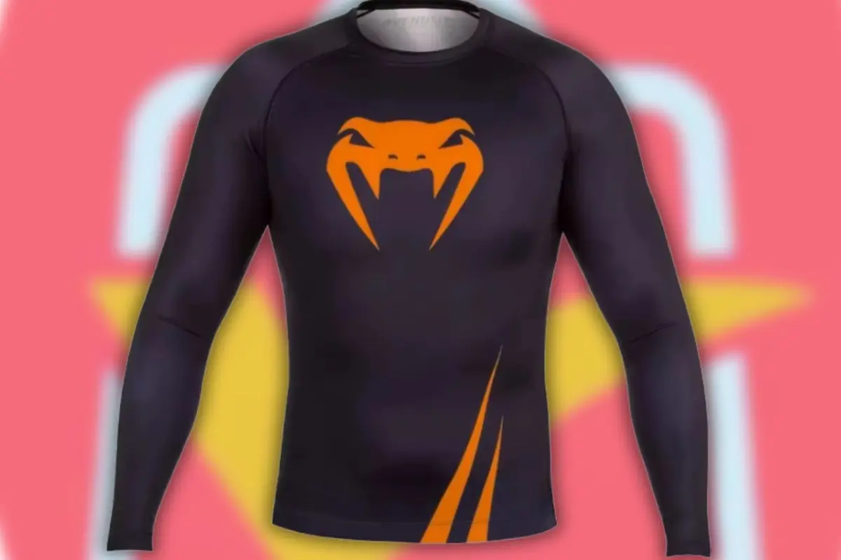 Playera deportiva negra de manga larga con logo naranja de serpiente estilizada en el pecho y líneas diagonales naranjas en la parte inferior sobre fondo rosa y amarillo desenfocado