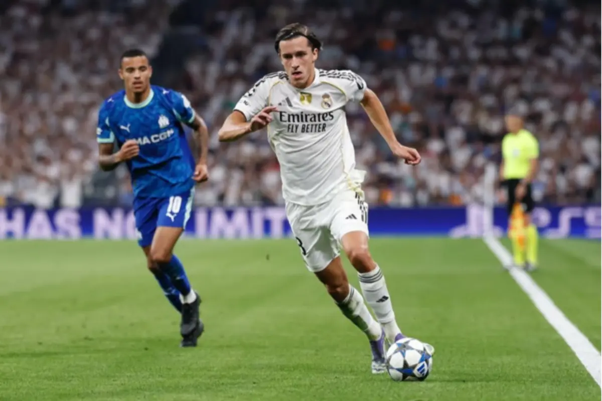 Futbolista del Reial Madrid amb uniforme blanc condueix la pilota en cursa per la banda mentre un rival amb samarreta blava el persegueix en un estadi ple de públic