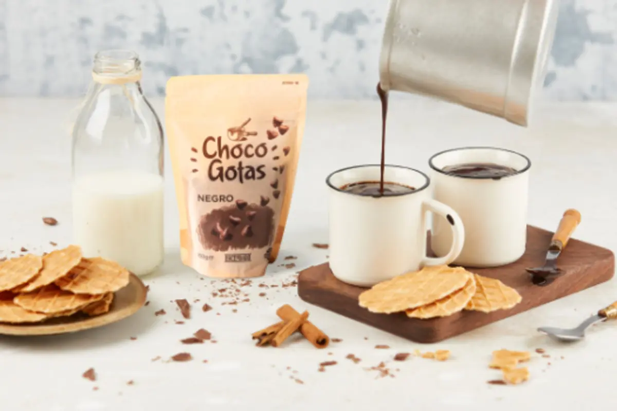 Taza de chocolate caliente servido con chispas de chocolate negro, galletas tipo oblea, canela y una botella de leche sobre una mesa clara