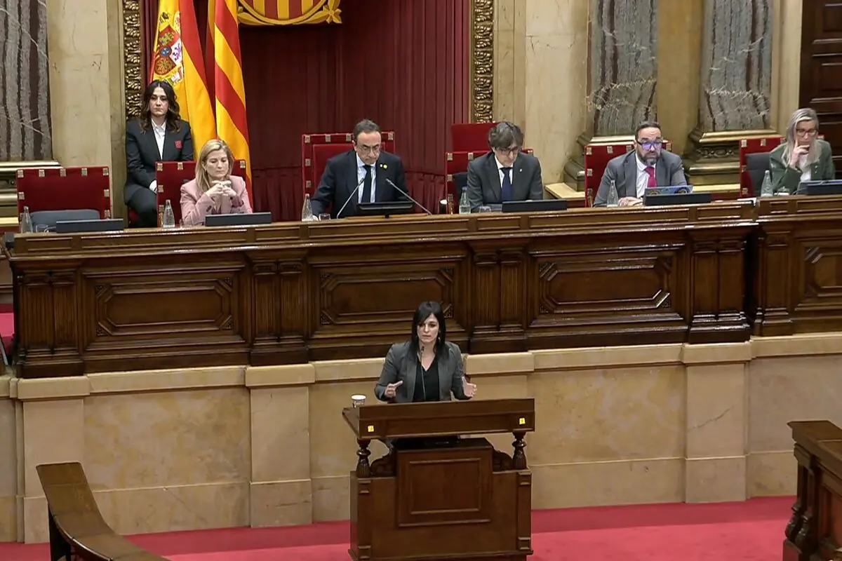 Mujer hablando en el atril de un parlamento con varios representantes sentados detrás en la mesa presidencial y banderas oficiales al fondo