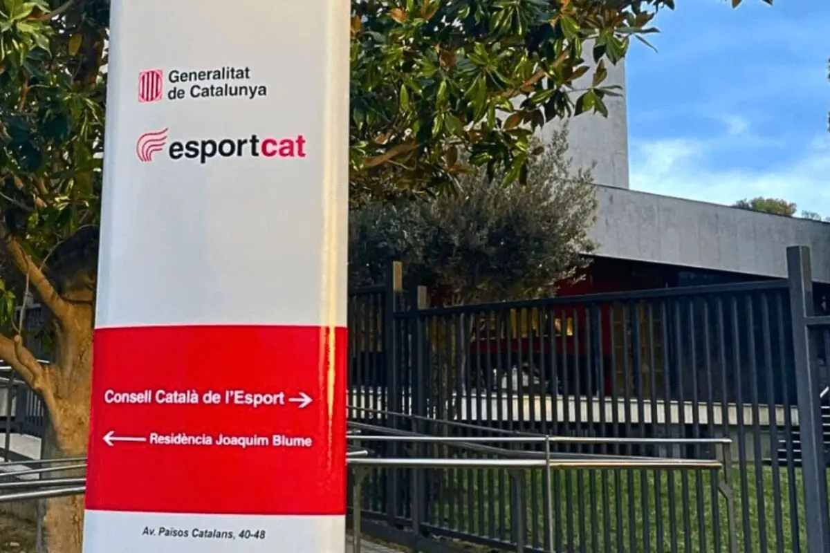 Tótem informativo de la Generalitat de Catalunya y esportcat frente a un edificio moderno con valla metálica y árboles en un entorno urbano
