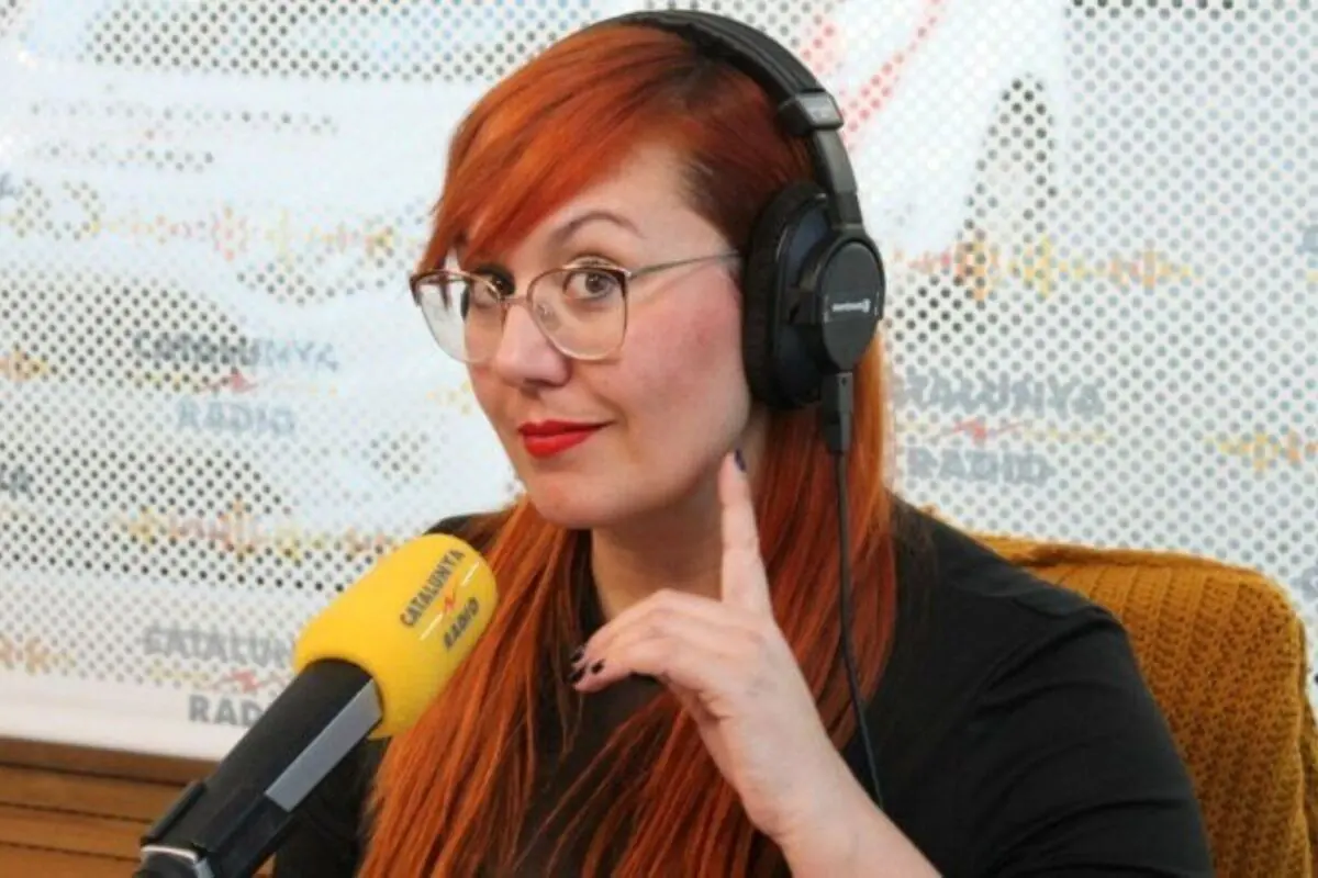 Dona pèl-roja amb ulleres i auriculars parlant davant d’un micròfon groc en un estudi de ràdio