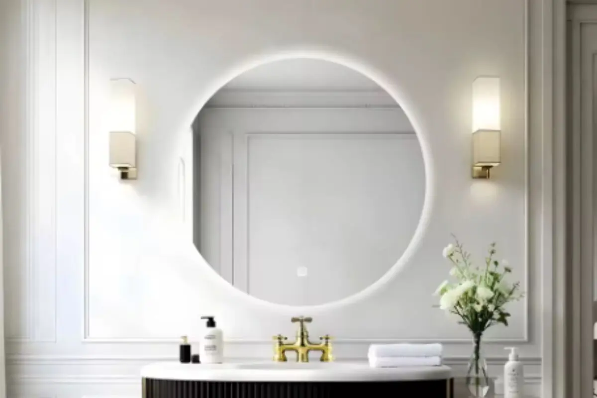 Baño moderno con espejo redondo retroiluminado grifería dorada y encimera blanca con flores y productos de tocador