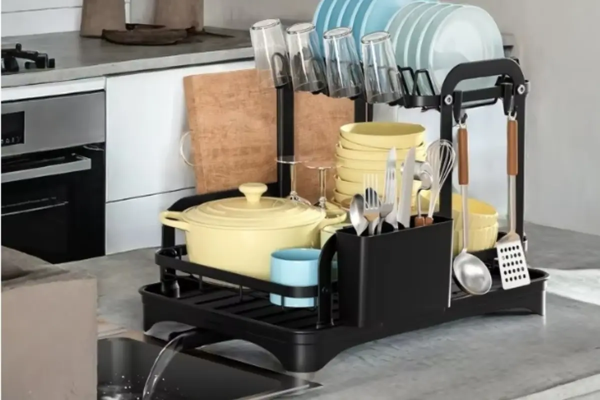 Escurridor de platos negro de dos niveles sobre encimera de cocina con vajilla, cubiertos y utensilios organizados y agua escurriendo hacia el fregadero