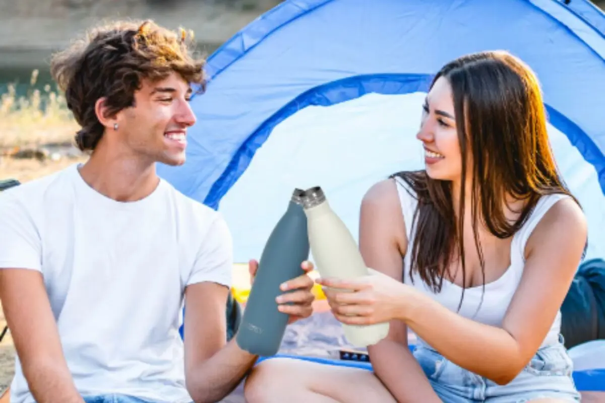 Pareja joven sentada frente a una tienda de campaña brindando con botellas reutilizables mientras sonríe al aire libre Pareja joven sentada frente a una tienda de campaña brindando con botellas reutilizables mientras sonríe al aire libre