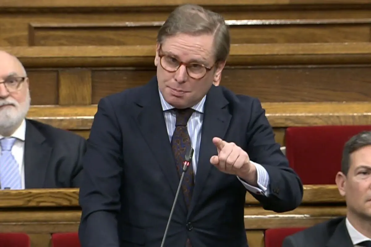 Hombre con gafas y traje oscuro hablando en un parlamento y señalando con el dedo mientras otros asistentes lo escuchan sentado detrás