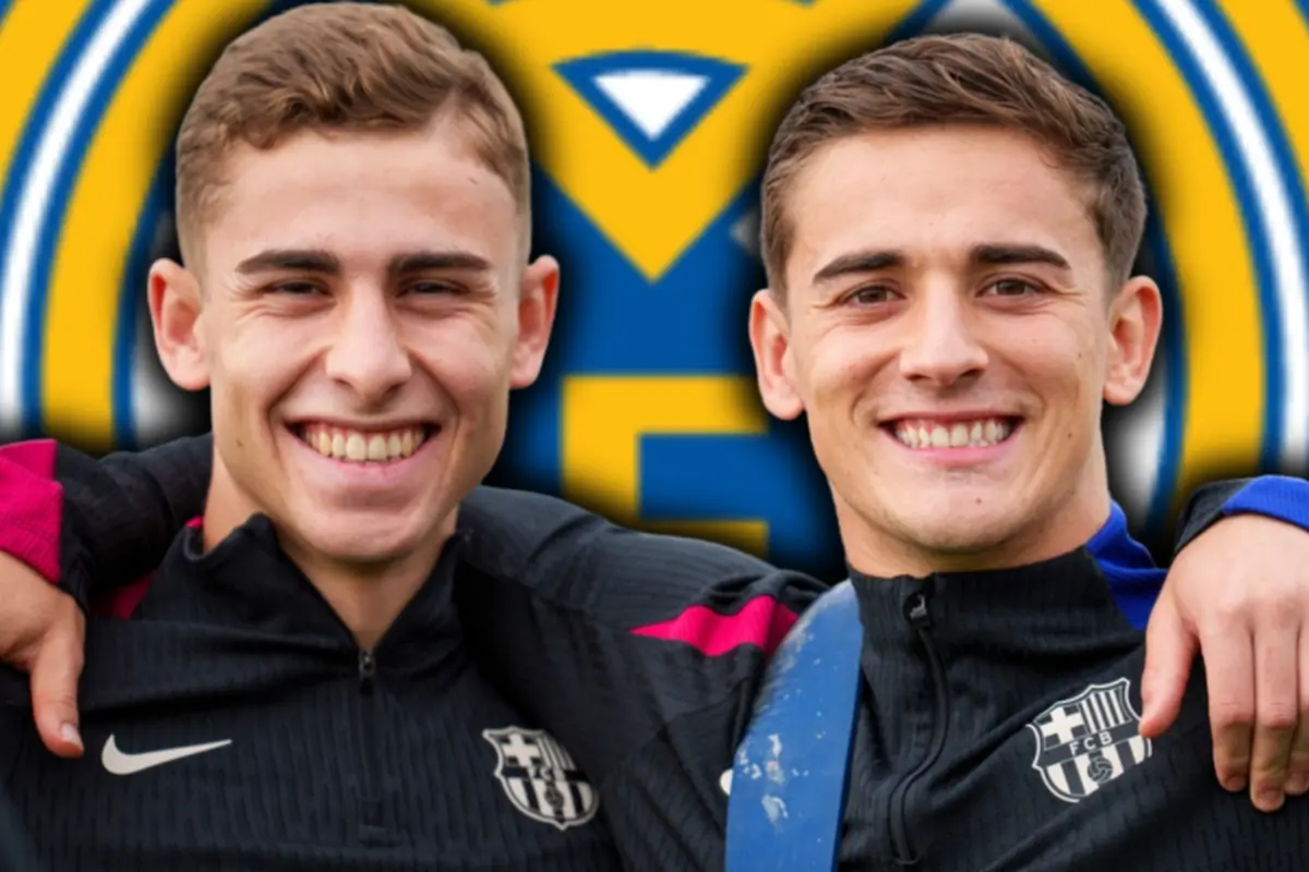 Dos joves futbolistes del FC Barcelona somriuen abraçats amb un gran escut blau i groc desenfocat de fons