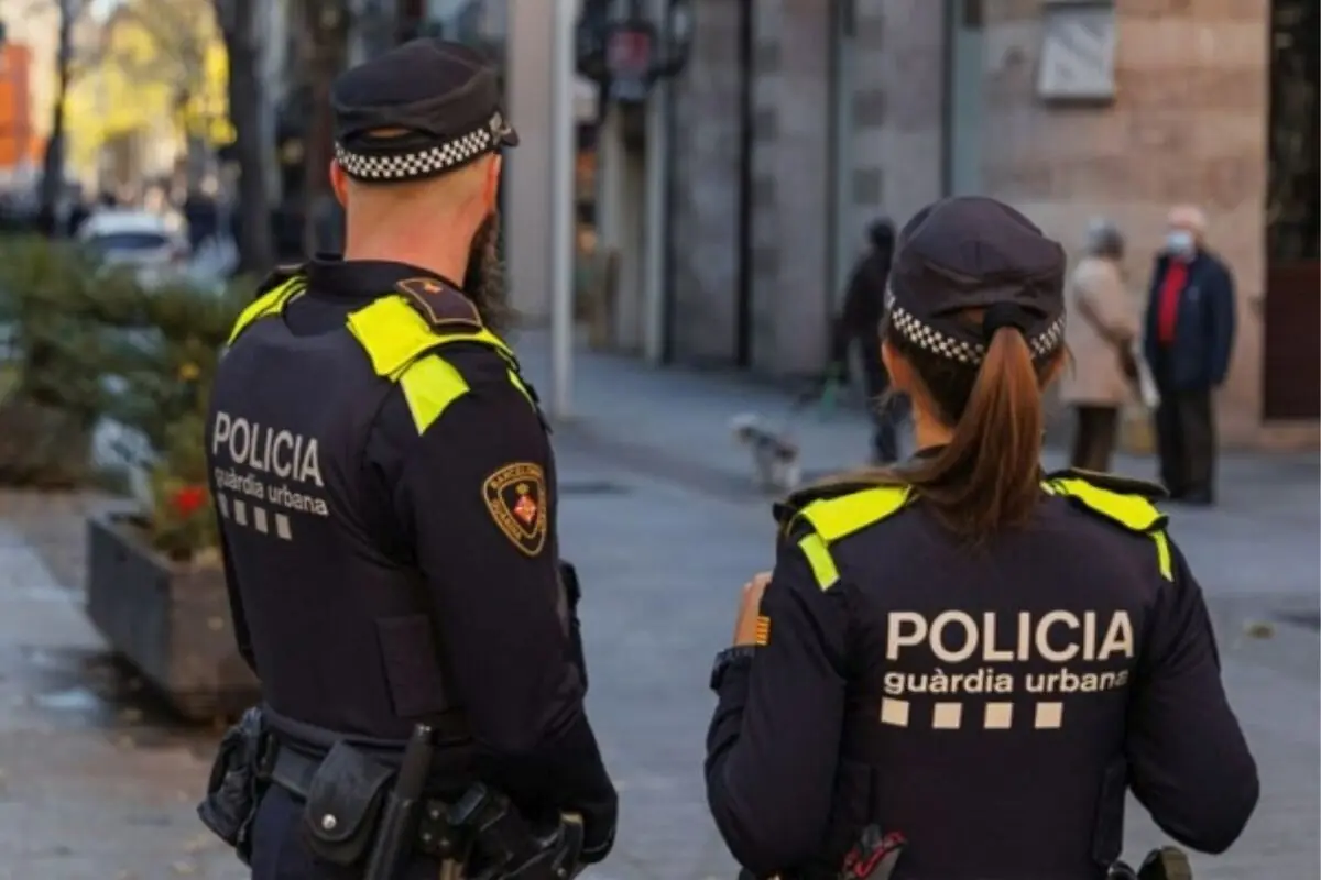 Dos agentes de la Guardia Urbana de espaldas patrullando una calle urbana con peatones al fondo