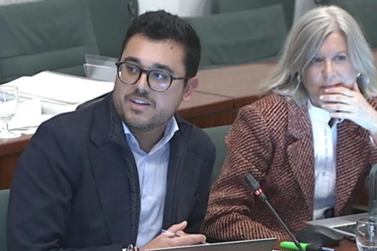 Home jove amb ulleres i jaqueta fosca parlant davant d’un micròfon al costat d’una dona de cabells canosos amb jaqueta marró en una sala de reunions