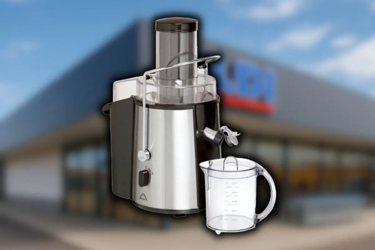 Extractor de jugos de acero inoxidable con vaso medidor transparente frente a la fachada borrosa de una tienda Aldi.