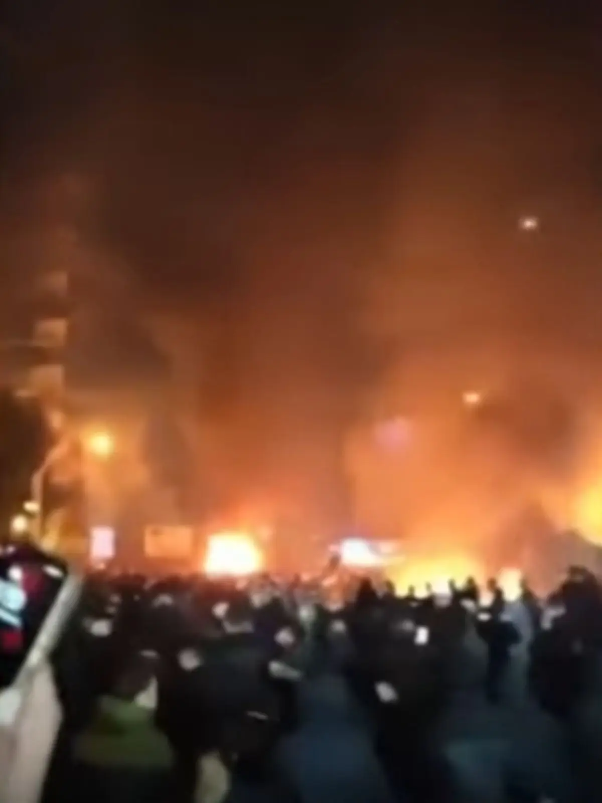 Multitud de persones reunides de nit davant de grans flames i fum en un carrer urbà de l’Iran.