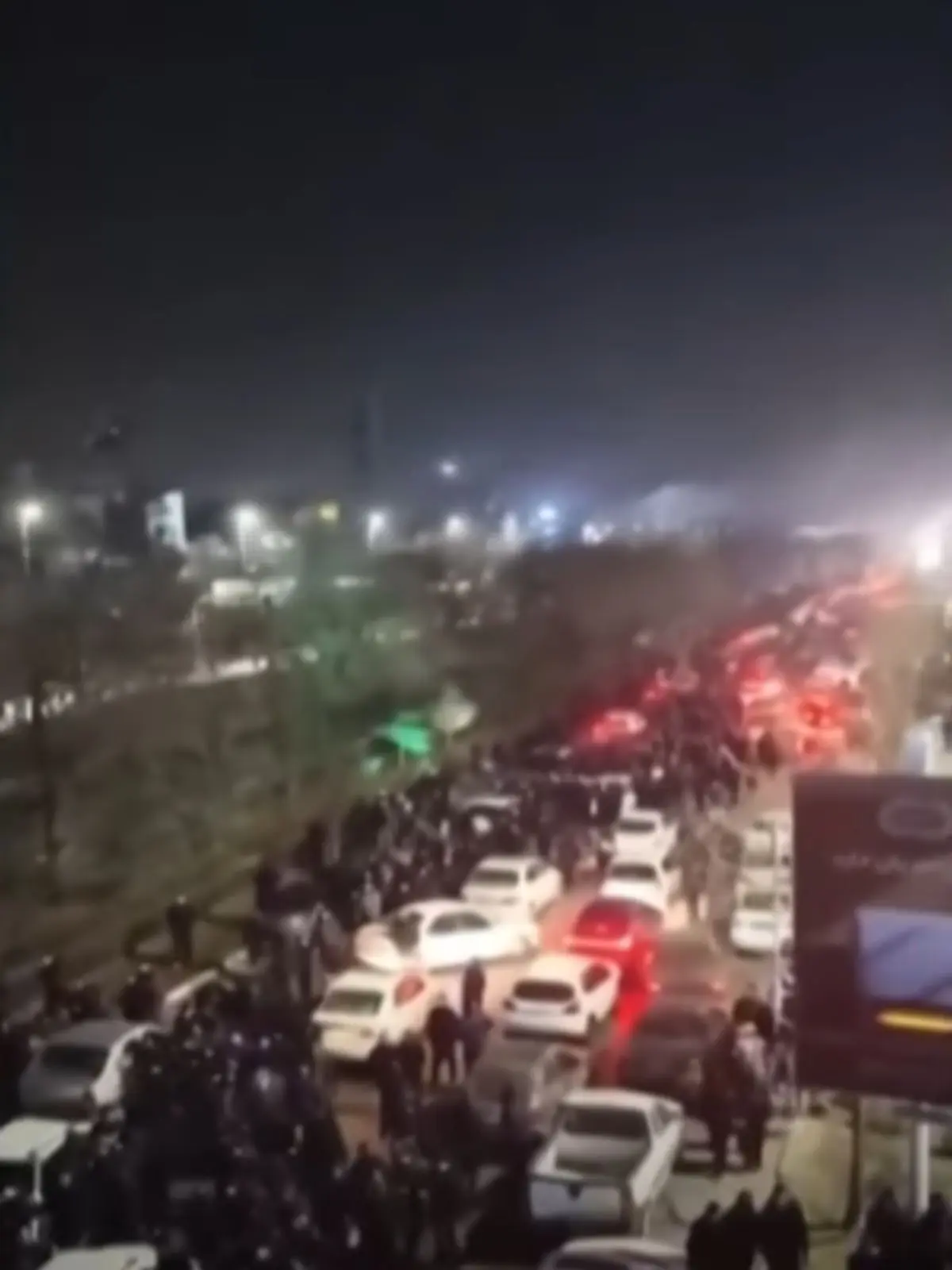Multitud de persones i vehicles omplen una avinguda urbana de nit, amb llargues fileres de cotxes i llums de fre vermelles il·luminant l’escena a l’Iran.