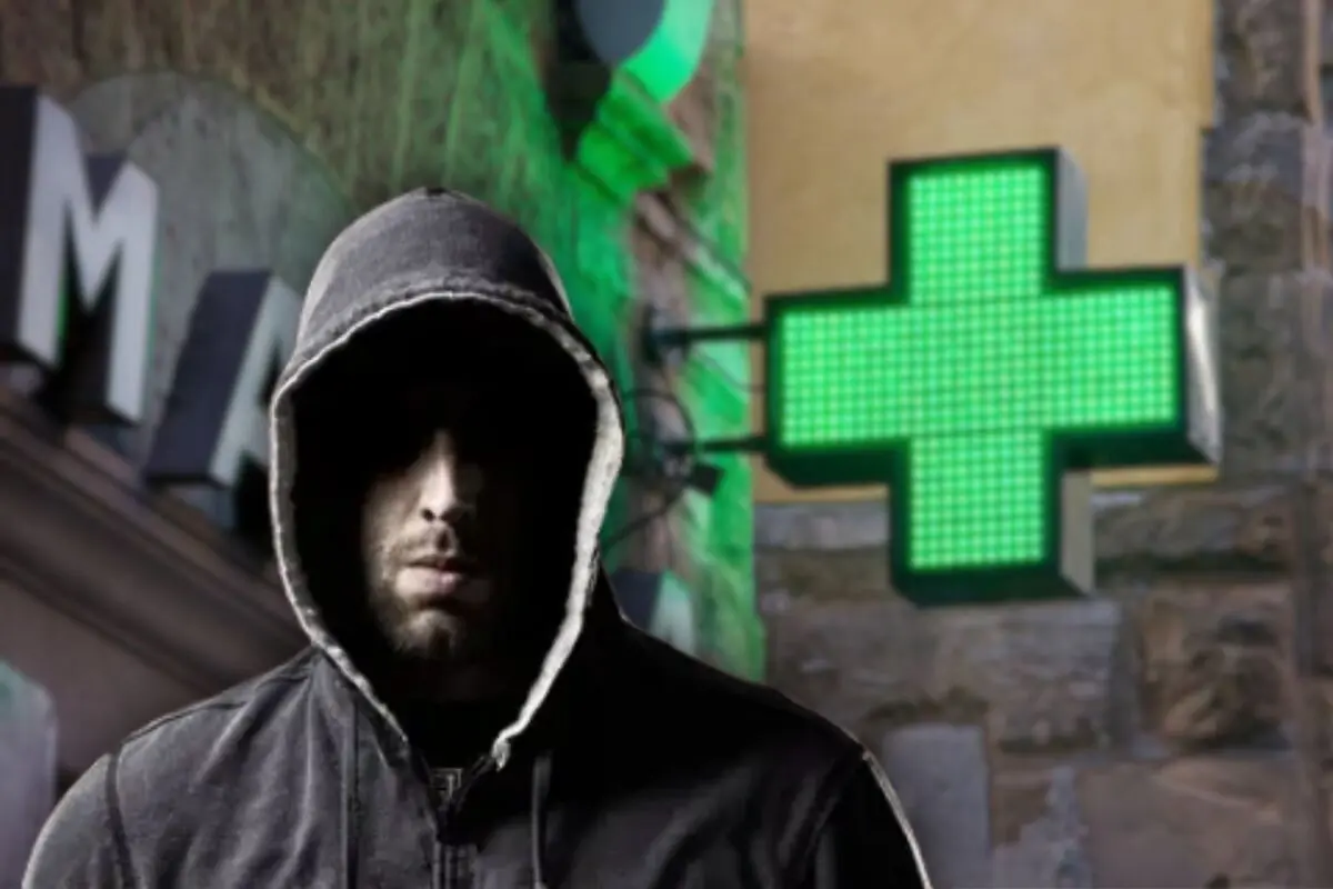 Hombre con sudadera y capucha oscura frente a un letrero luminoso verde de farmacia