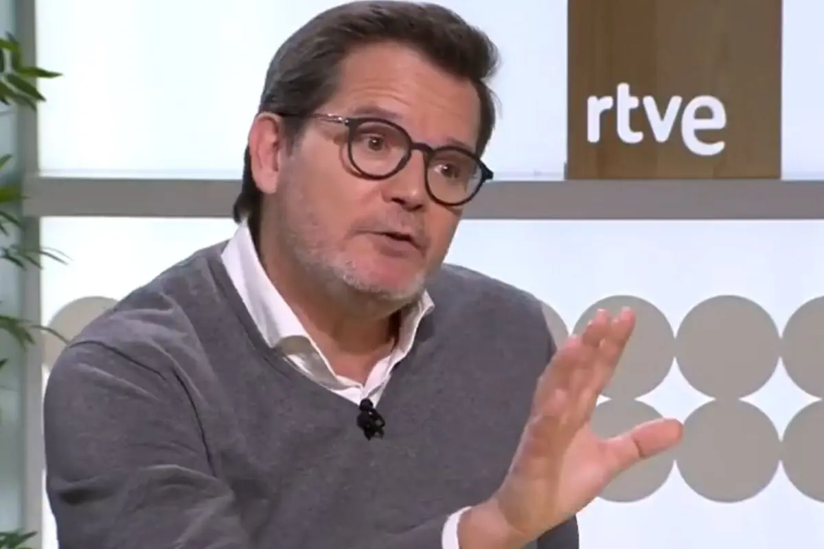 Hombre con gafas y jersey gris hablando gesticulando con una mano en un plató de televisión con el logo de rtve al fondo