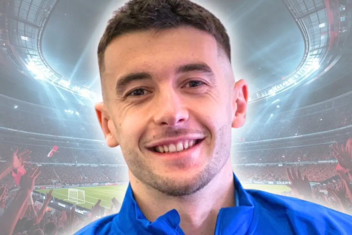 Retrat d’un futbolista somrient amb jaqueta blava sobre un estadi ple d’aficionats i llums brillants