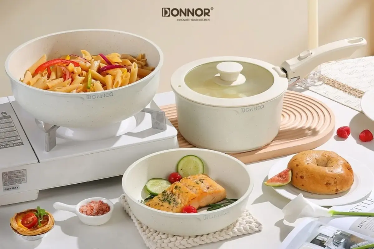 Juego de sartenes color blanco con pasta, salmón y pan bagel servidos en una mesa de cocina iluminada y decorada con frutas y accesorios de mesa