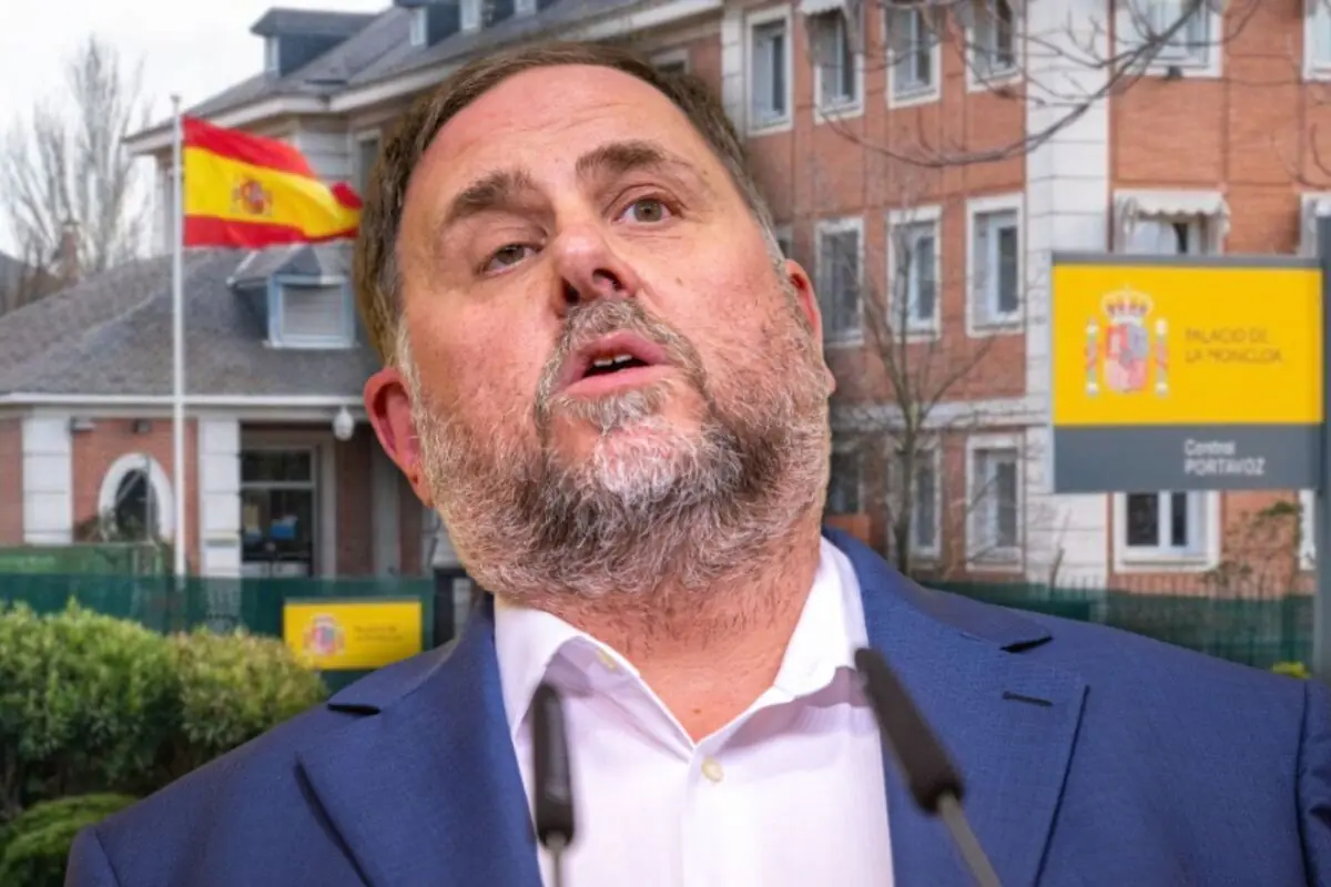 Hombre de mediana edad con barba hablando ante micrófonos con un edificio gubernamental español y una bandera de España al fondo