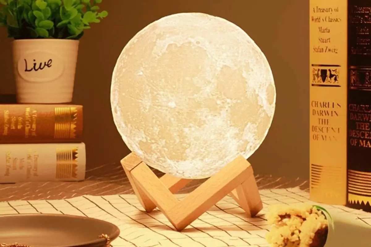 Lámpara decorativa con forma de luna iluminada sobre base de madera en una mesa junto a libros y una maceta con planta Lámpara decorativa con forma de luna iluminada sobre base de madera en una mesa junto a libros y una maceta con planta